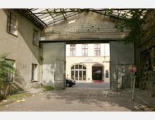 Ein Kopfsteinpflaster-Hof mit einem offenen Metalltor führt zu einem hellen Gebäude mit der Aufschrift "Hotel Augustinenhof". Das Gebäude hat große Fenster und einen Eingang mit roten Teppichvorleger; links und rechts vom Tor befinden sich ältere, verwitterte Mauern.