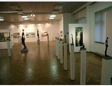 Ein Ausstellungsraum mit Holzboden und weißen Wänden, in dem Skulpturen auf weißen Säulen sowie Gemälde an den Wänden ausgestellt sind. Die Decke ist mit rechteckigen Leuchten versehen, und große Fenster lassen natürliches Licht herein.