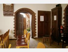 Innenraum mit hellen Holzstühlen auf einem Holzboden, einer offenen Tür mit gemauertem Rundbogen aus roten Ziegelsteinen und Schränken mit dunklen Holztüren. Im Hintergrund ist ein Bereich mit weiteren Ziegelsteinen und einem Blumenarrangement sichtbar.