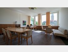 Heller Besprechungsraum mit Holzmöbeln, darunter ein großer rechteckiger Tisch mit gepolsterten Stühlen und ein Schreibtischbereich vor drei großen Fenstern. Durch die Fenster ist ein historisches Gebäude mit Turm auf der gegenüberliegenden Straßenseite zu sehen.