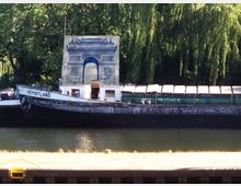 Ein Frachtschiff mit der Aufschrift "HEIMATLAND" liegt in einem Fluss am Ufer, umgeben von Bäumen und Grün. Auf dem Schiff ist ein ungewöhnlich montierter, kunstvoll verzierter Triumphbogen zu sehen.