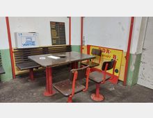 Holztisch mit festmontierten roten Metallstühlen und einer Holzbank an einer Wand in einem Raum mit abgenutztem Boden und grün-weiß gestrichenen Wänden. Im Hintergrund befinden sich eine alte Namensliste, eine DDR-Werbetafel und ein Karton mit der Aufschrift "VERITAS".