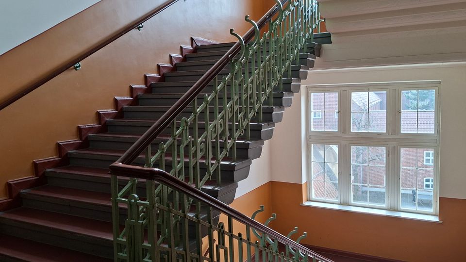 Innenansicht eines Treppenhauses mit breiter, dunkel gestrichener Holztreppe und grünem Metallgeländer mit Holzhandlauf, die in einer Kurve nach oben führt. Rechts befindet sich ein großes, mehrteiliges Fenster, durch das Ziegeldächer und Gebäude im Außenbereich sichtbar sind.