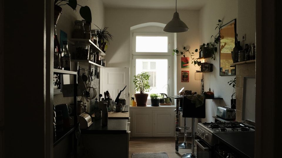 Schmale Wohnküche mit hohen weißen Wänden und großem Fenster am Ende, dessen Fensterbank mit Topfpflanzen und Kräutern bestückt ist. Links und rechts stehen dunkle Arbeitsflächen, offene Regale mit Gläsern und Küchenutensilien sowie ein Gasherd; eine Hängelampe und Wanddekorationen ergänzen den Raum.