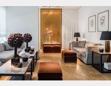 Eleganter Wohnraum mit beigen Marmorböden, grauen Sofas, braunen Samthockern und schwarzen Beistelltischen, darauf dekorative Blumenarrangements. Im Hintergrund eine goldene Wandverkleidung, flankiert von minimalistisch gestalteten Kunstwerken und schlanken Metallskulpturen.