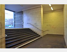 Eingangsbereich einer U-Bahn-Station mit Treppen, die zu einer geöffneten Gittertür führen, umgeben von Wänden mit beigefarbenen Fliesen. Neonröhren beleuchten den Raum, während durch die Tür ein Blick auf eine Straße mit Bäumen und Gebäuden sichtbar ist.