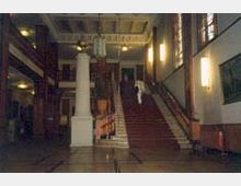 Großes, historisches Treppenhaus mit roten Teppichstufen, flankiert von weißen Säulen und Geländern. Der Raum hat hohe Decken mit Leuchtern, große Fenster an der rechten Wand und Wandlampen, die weiches Licht spenden.