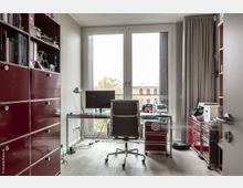 Arbeitszimmer mit modernem Design, ausgestattet mit einem Glastisch, einem Schreibtischstuhl und roten Metallregalen an der linken und rechten Wand. Der Raum verfügt über ein großes Fenster mit Blick auf Bäume und ein historisches Gebäude im Hintergrund.
