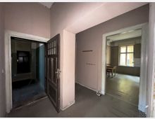 Ein leerer, heller Innenraum mit schlichten Wänden; links eine geöffnete hölzerne Zimmertür führt in einen dunkleren Raum, während rechts ein Durchgang in einen Raum mit einem Fenster führt, durch das Tageslicht einfällt. Im rechten Raum stehen ein Tisch und ein Stuhl vor dem Fenster.