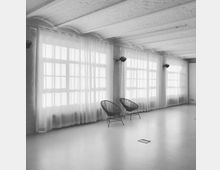 Heller Raum mit großen Fenstern, die von transparenten Vorhängen bedeckt sind, und einer Decke mit sichtbaren Trägern. Im Vordergrund stehen zwei schlichte Stühle mit Drahtgestell auf einem glatten Boden.