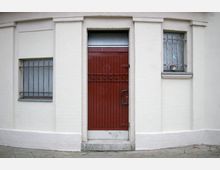 Beigefarbene Hausfassade mit einer dunkelroten Tür, die mit dem Schriftzug "Herren" versehen ist, flankiert von zwei kleinen Fenstern mit Metallgittern. Der Boden davor ist gepflastert.