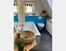 Ein Badezimmer mit Dachschräge, einer freistehenden Naturstein-Waschschale auf einem Holztisch, einer Badewanne mit Glasspritzschutz und blauen Fliesen im Fischgrätmuster an der Wand. Ein Fenster mit bunter Musterscheibe und gelben Vorhängen sowie eine Toilette und ein Heizkörper sind ebenfalls sichtbar.