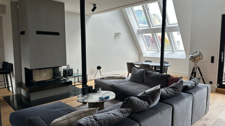 Modernes Dachgeschoss-Wohnzimmer mit großer grauer Sofalandschaft, rundem Couchtisch und freistehendem Kamin auf schwarzer Steinplatte. Schräge Decke mit großen Dachfenstern und Blick nach draußen, dahinter ein Esstischbereich sowie ein Stativ-Scheinwerfer neben einer Glastür.