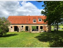 Ein Bauernhaus aus Naturstein und Backstein mit roten Dachziegeln, umgeben von einer grünen Rasenfläche. Links blüht ein Fliederbusch, rechts stehen schattenspendende Bäume, im Hintergrund ist eine ländliche Landschaft mit Feldern und einem Gewässer sichtbar.