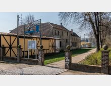 Eingangsbereich eines Sportgeländes mit einem Schild „Ernst Stadion 1977“, flankiert von Backsteinsäulen. Im Hintergrund sind ein Fachwerkgebäude, ein älteres Gebäude mit verblasster Fassade und ein gelbes Gebäude mit Graffiti zu sehen, umgeben von Bäumen und einem Weg.