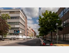 Straßenszene in einem städtischen Gewerbegebiet mit modernen mehrstöckigen Gebäuden aus Glas und Metallfassaden. Links und rechts der Straße befinden sich geparkte Autos und vereinzelte Bäume, darüber ein leicht bewölkter Himmel.