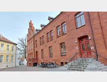 Backsteingebäude mit großen Fenstern und einem spitzgiebligen Turmelement auf der linken Seite, davor eine Reihe abgestellter Fahrräder; rechts führt eine Treppe mit Geländer zu einer roten Holztür. Links im Bild steht ein gelbes Gebäude mit weißen Fensterrahmen.