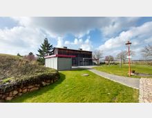 Ein moderner Außenbereich mit einem Holzgebäude und einem kleinen, grauen Nebengebäude, umgeben von grünem Rasen, Bäumen und einem gepflasterten Weg. Im Hintergrund sind offene Felder und ein teilweise bewölkter Himmel sichtbar.