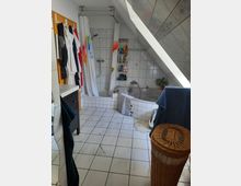 Badezimmer mit weiß gefliesten Wänden und Böden, einer Badewanne mit Duschvorhang unter einer Dachschräge. An der Wand befinden sich Regale mit Pflegeprodukten, und ein Wäschekorb aus Holzgeflecht steht im Vordergrund.