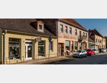 Straßenzug in einer Kleinstadt mit niedrigen, aneinandergebauten Wohn- und Geschäftshäusern mit Ziegeldächern und Ladenfronten, darunter ein Optiker und ein älteres Geschäft mit verblasster Fassadenaufschrift. Entlang der Straße stehen mehrere geparkte Autos, davor verläuft ein schmaler Gehweg mit Kopfsteinpflaster und Geländern.