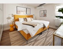 Modernes Schlafzimmer mit einem gelben Polsterbett, weißen Bettwäsche und Kissen in neutralen Farben. Der Raum besitzt weiße Holzvertäfelung, einen Teppich mit Blättermuster, hängende Wandbilder, eine Klimaanlage und ein Fenster, das Tageslicht hereinlässt.