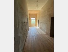 Ein langer, schmaler Flur mit rohen, unverputzten Wänden und einem Holzfußboden. Am Ende des Flurs befindet sich eine verglaste Doppeltür, durch die Tageslicht einfällt, sowie offene Kabel an der Decke.