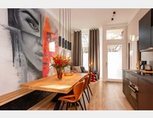 Modernes Wohnküchen- und Esszimmer mit großem Holztisch und Lederstühlen, schwarzer Einbauküche und hängenden Zylinder-Pendelleuchten. An der linken Wand befindet sich ein großformatiges Wandbild, am Ende des Raums eine hohe Glas-/Terrassentür mit Vorhängen und viel Tageslicht.