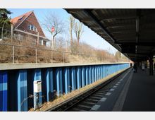 Bahnhof mit überdachtem Bahnsteig und Gleis, daneben eine blaue Stützmauer und dahinter eine Böschung mit Geländer. Im Hintergrund ist ein Backsteingebäude mit spitzem Giebel sowie spärlicher Vegetation zu sehen.