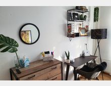 Ein kleiner, gemütlicher Arbeitsbereich mit weißer Wand, an der ein rundes Spiegel und ein Metall-Regal mit Dekoobjekten und Büchern hängt. Ein hölzernes Sideboard steht links, daneben ein dunkler Schreibtisch mit Pflanze, Büromaterial und geflochtenem Stuhl; rechts eine Stehlampe.