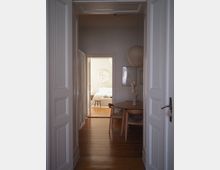 View through white paneled double doors into a narrow apartment hallway with wooden floors, leading to a bright bedroom. A small dining nook with a round table, chairs, framed wall art, and a hanging paper lantern sits on the right, with a curtain along the left wall.