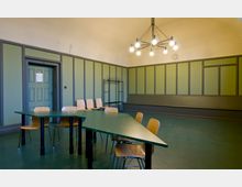 Raum mit grün gestrichenen Wänden und einer Sitzgelegenheit aus Holzbänken und Stühlen um grüne Tische. An der Decke hängt ein moderner Kronleuchter, an der Wand ist eine Garderobe und eine Uhr angebracht.