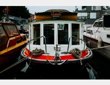Ein kleines Motorboot mit rotem und weißem Bug, festgemacht zwischen anderen Booten in einem Hafen. Im Hintergrund sind Gebäude und Bäume sichtbar.
