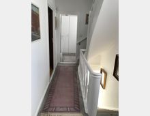 Schmaler, heller Flur in einem Wohnhaus mit weiß gestrichenen Wänden und einem violetten, gemusterten Teppichläufer. Rechts befindet sich ein Treppenabsatz mit weißem Geländer, links sind Türen und Wandbilder, am Ende des Flurs steht ein weißer Einbauschrank.