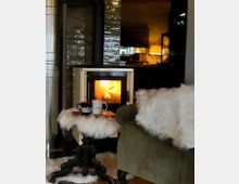 Gemütliche Inneneinrichtung mit einem brennenden Kaminofen vor einer glänzenden, dunklen Fliesenwand. Ein grüner Sessel mit weißem Fellüberwurf und ein kleiner Tisch mit Fellauflage und Tassendekoration stehen im Vordergrund, während eine warme Beleuchtung den Hintergrund erhellt.