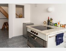 Kleine, helle Küche mit gemusterten Fliesen auf dem Boden, einer Arbeitsplatte aus Marmor mit eingebautem Kochfeld und Ofen, einem kleinen Kühlschrank an der Wand und verschiedenen Gewürzen sowie Flaschen darauf. Im Hintergrund ist eine Holztreppe und ein Regal mit einem Korb erkennbar.