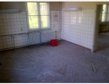 Ein leerer Raum mit weißen gefliesten Wänden, Holzboden im Fischgrätmuster und einem großen Fenster mit Blick ins Grüne. Unter dem Fenster befindet sich ein Heizkörper, und auf dem Boden steht eine rote Kiste.