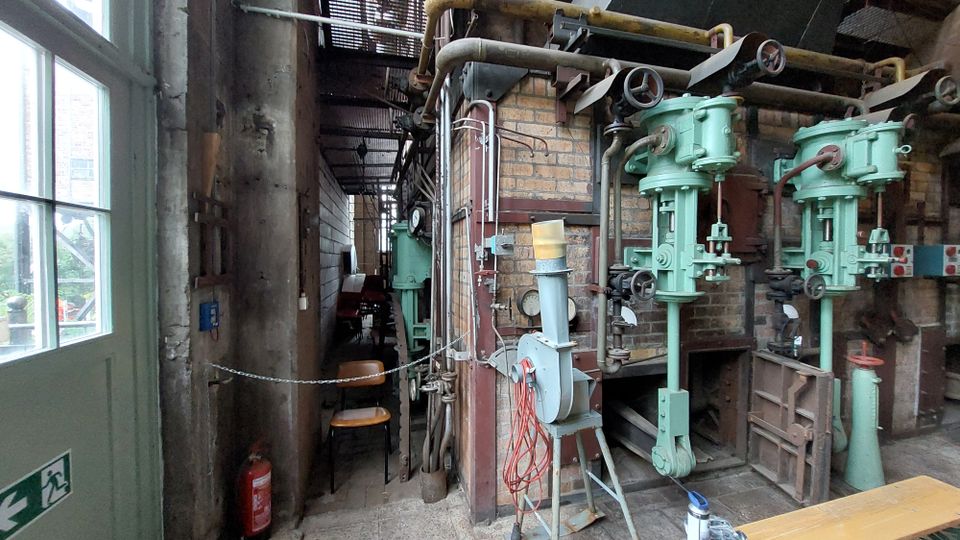 Industrieanlage mit Backsteinwänden und grünen, massiven Maschinenkomponenten, darunter Rohre und Ventile, die an einer Wand montiert sind. Im Vordergrund eine Tür mit Glaseinsätzen und ein Feuerlöscher; im Hintergrund eine Reihe von Stühlen und schmales Innenraumlicht.