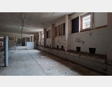 Langer, verlassener Innenraum in einem Betongebäude mit niedriger Decke, Stützpfeilern und einer Reihe kleiner, vergitterter Fenster entlang der rechten Wand. Die Wände sind mit Graffiti beschmiert, der Boden ist staubig und mit Schutt bedeckt, am Ende des Gangs sind Gittertüren und abgetrennte Bereiche zu sehen.