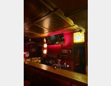 Innenraum einer Bar mit langem Holzthekenbereich, roten Wänden und Regalen mit Flaschen und Gläsern. An der Decke sind dekorative Holzpaneele zu sehen, daneben Spiegel, runde Wandleuchten und ein beleuchtetes Schild mit Text.