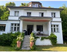 Weiße zweistöckige Villa mit Satteldach und weiß gerahmten Fenstern, umgeben von Bäumen. Im Vordergrund eine überdachte Veranda mit eingefasster Metallbrüstung, blühenden Pflanzen und einer kurzen Treppe, die zu einer Rasenfläche führt.