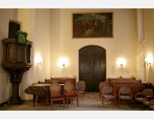 Innenraum einer Kirche mit einer dunkelbraunen hölzernen Kanzel links, einer geschlossenen Doppeltür in der Mitte und mehreren Stühlen vor hölzernen Kirchenbänken. Über der Tür hängt ein großes Gemälde mit einer religiösen Szene, und an den Wänden befinden sich Wandlampen, die warmes Licht spenden.