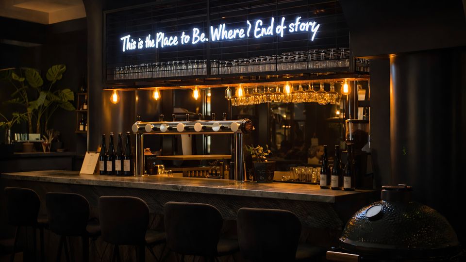 Moderne Bar mit langem Tresen und Barhockern, darüber hängende Weingläser und warm leuchtende Edison-Glühbirnen. Im Hintergrund sind Flaschenregale und ein Neon-Schriftzug an einer dunklen Wandverkleidung zu sehen.