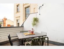 Kleine Terrasse mit Holzfußboden, einem quadratischen Tisch und vier passenden Stühlen. An der weißen Wand hängt eine grüne Pflanze, daneben verläuft eine Lichterkette, und im Hintergrund ist die Fassade eines hellen Altbaus zu sehen.