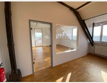 Ein heller Büroraum mit hellem Parkettboden, großen Fenstern und Glaswänden, die in einen angrenzenden Raum blicken. Sichtbare schwarze Stahlträger an den Wänden und eine Feuertreppe im linken Bildbereich geben einen industriellen Charakter.