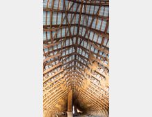 Holzstruktur eines großen Dachbodens mit sichtbaren Dachbalken und -streben, gedeckt mit Wellblechplatten. Trockenes Stroh hängt in Bündeln von den Holzbalken herab, und der Boden ist teilweise mit Heu bedeckt.