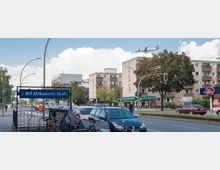 Straßenszene in Berlin mit dem U-Bahneingang "U-Bhf Afrikanische Straße" im Vordergrund, umgeben von geparkten Autos, Fahrradständern und Wohngebäuden. Im Hintergrund fliegt ein Flugzeug über mehrstöckige Wohnhäuser und Bäume, während eine Straße mit Ampeln die Szene durchzieht.