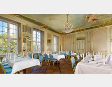 Eleganter Speisesaal mit weißen Wänden, goldenen Verzierungen und großen Fenstern, die Tageslicht einlassen. Der Raum ist mit einer bemalten Decke, einem opulenten Kronleuchter und weiß gedeckten Tischen mit blauen Stühlen ausgestattet.