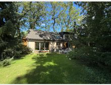 Einfamilienhaus mit Holzfassade und dunklem Schrägdach, umgeben von hohen Bäumen und einem grünen Rasen. Im Vordergrund befindet sich eine Terrasse mit Gartenmöbeln, im Hintergrund dichter Wald.