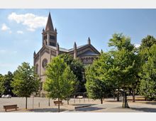 Backsteinkirche mit einem hohen Turm und neugotischen Architekturdetails, umgeben von Bäumen und einer weitläufigen, gepflasterten Platzfläche, auf der vereinzelt Bänke stehen. Der Himmel ist klar und blau.
