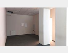 Ein leerer Innenraum mit weißen Wänden, grauem Teppichboden und abgehängter Decke mit eingebauten Leuchtplatten. An der linken Wand befindet sich ein Heizkörper, an der Rückwand sind zwei weiße Paneele und ein kleiner gefliester Bereich angebracht. Ein Teil des Raumes ist durch eine weiße Säule abgetrennt.